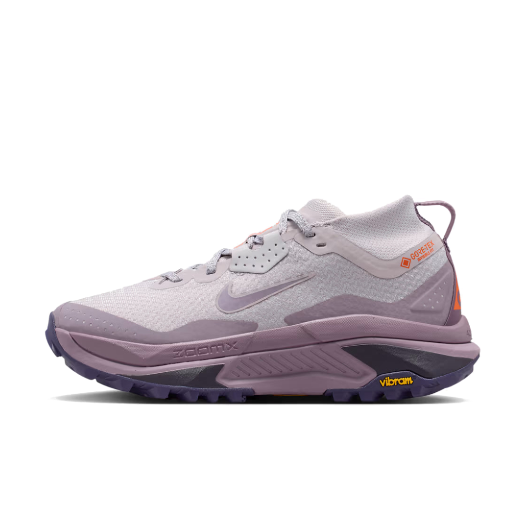 A Nike ACG ZEGAMA ZOOMX trail running shoe in a light lavender and purple colorway, featuring a textured mesh upper, a purple Swoosh, and a purple Vibram outsole, shown from a side profile on a light gray background.