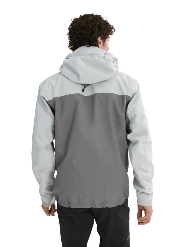 A man viewed from behind wearing a color-blocked hooded jacket with a light gray upper section and sleeves, and a darker gray lower back panel, paired with black pants against a white background. - Arc'teryx Beta AR GORE-TEX Jacket (Men's) - Solitude/Void