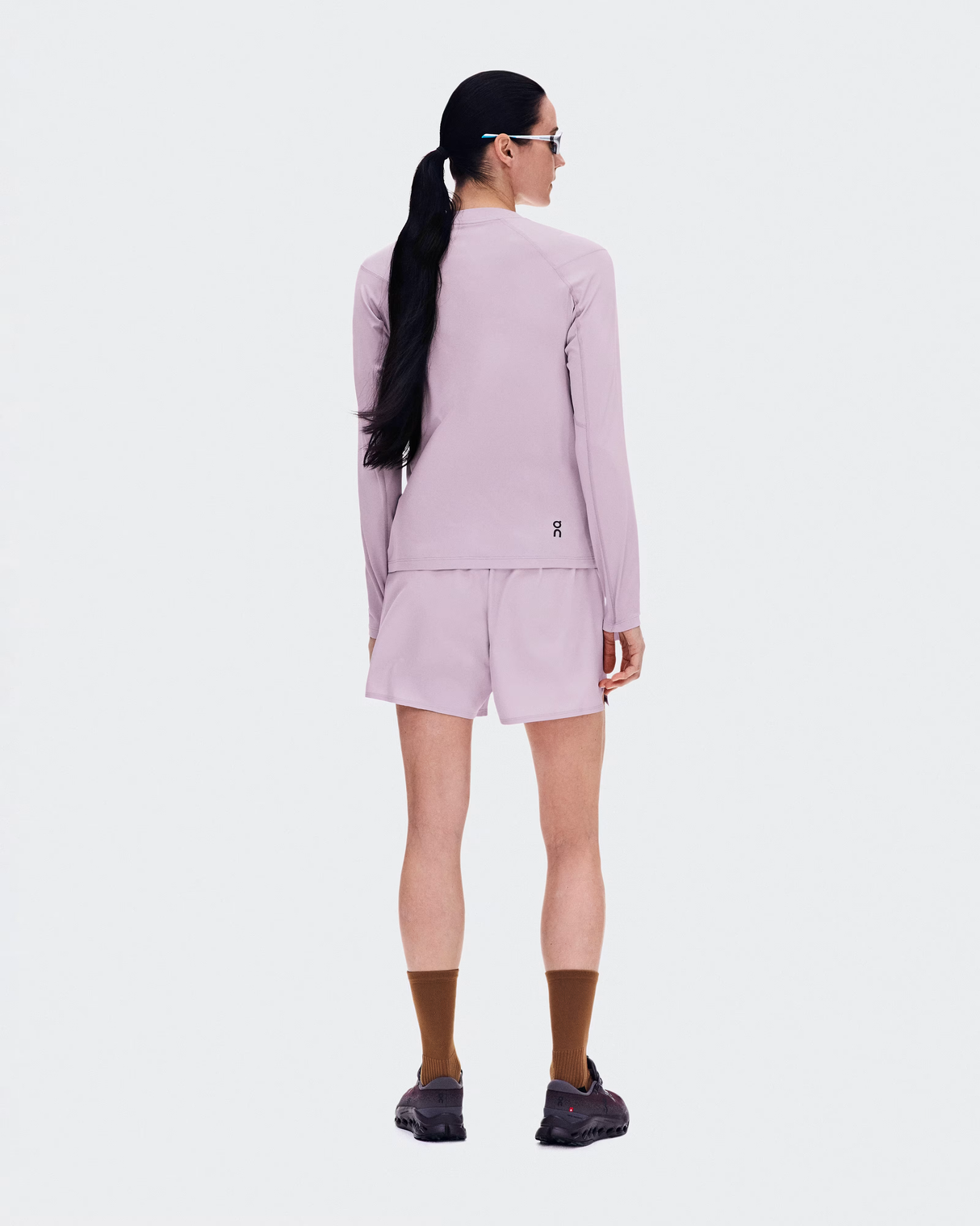 A model shown from the back wearing a matching light purple athletic set consisting of a long-sleeve zip-up jacket and shorts, paired with brown socks and dark sneakers against a light gray background. - On Trail Shorts (Women's) - Foxglove