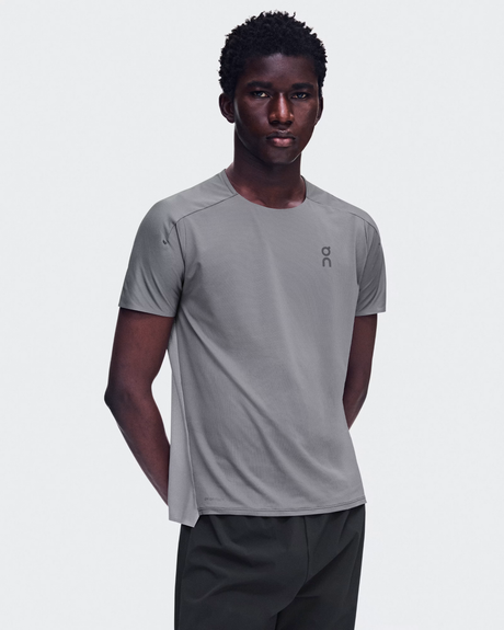 A man wearing a light gray athletic t-shirt with a small dark gray 'on' logo on the left chest, paired with dark gray athletic pants, against a light gray studio background. - On Performance-T (Men's) - Ember/Ore