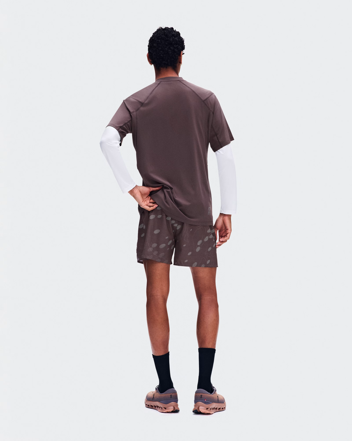 A man shown from the back wearing a brown short-sleeve t-shirt layered over a white long-sleeve shirt, matching brown shorts with a subtle pattern, black socks, and brown sneakers, against a light gray background. - On Trail-T Solar (Men's) - Mulberry
