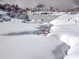 A snowy winter landscape with deep snowbanks, a partially frozen stream, snow-covered trees, and distant mountains under a cloudy sky. - Rob Blakers Wild Tasmania 2027 Large Calendar