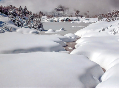 A snowy winter landscape with deep snowbanks, a partially frozen stream, snow-covered trees, and distant mountains under a cloudy sky. - Rob Blakers Wild Tasmania 2027 Large Calendar