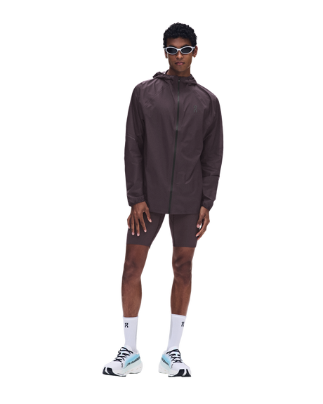 A male model wearing a dark brown athletic windbreaker jacket, matching shorts, white socks, and white sneakers with light blue accents, standing against a solid white background. - On Ultra Jacket (Men's) - Urchin