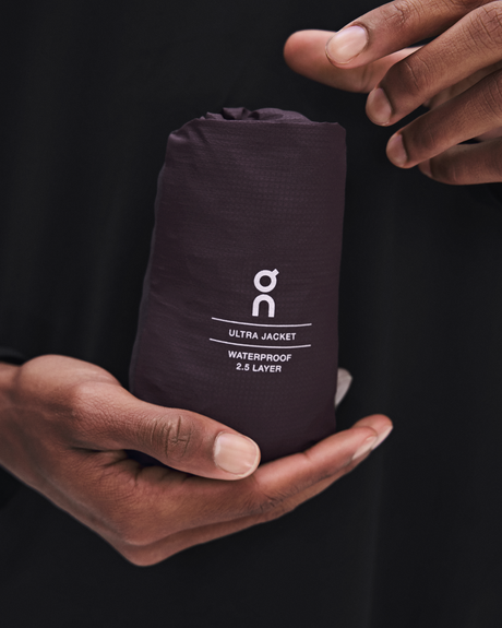 A close-up of a person holding a dark purple, compactly packed On Ultra Jacket. The person's hands are visible holding the pack, and the background is a solid black fabric. - On Ultra Jacket (Men's) - Urchin