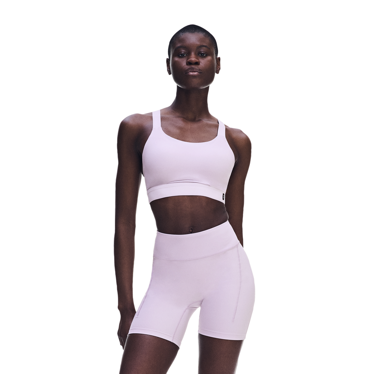 A model wearing a light pink matching activewear set consisting of a sports bra and high-waisted biker shorts, photographed against a solid white background. - On Performance Bra (Women's) - Foxglove