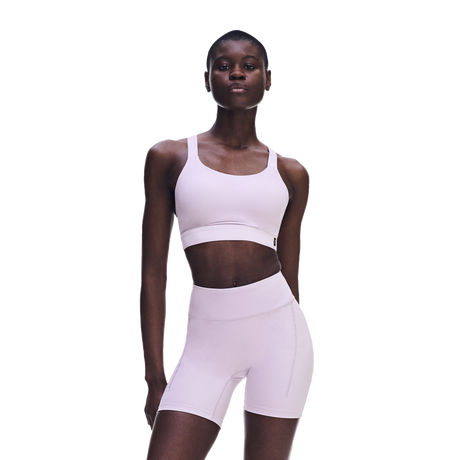 A model wearing a light pink matching activewear set consisting of a sports bra and high-waisted biker shorts, photographed against a solid white background. - On Performance Bra (Women's) - Foxglove