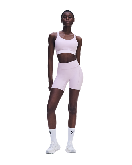 A model wearing a light pink athletic set consisting of a sports bra and matching high-waisted biker shorts, paired with white socks and white sneakers, set against a solid white background. - On Performance Bra (Women's) - Foxglove