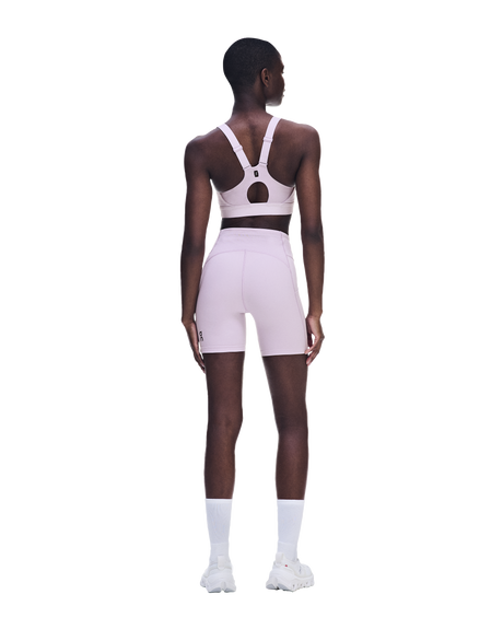 A model shown from the back wearing a light pink athletic set consisting of a sports bra with an open back and matching high-waisted shorts, paired with white socks and white sneakers on a solid white background. - On Performance Bra (Women's) - Foxglove