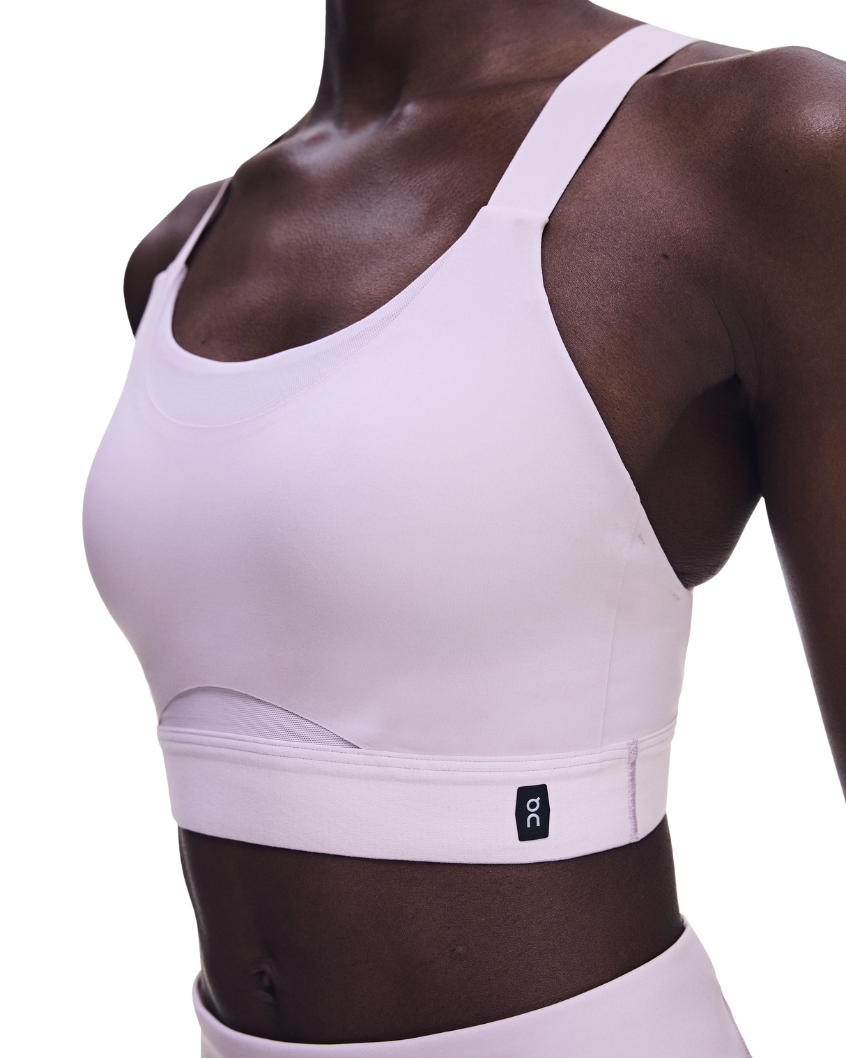 A close-up of a model wearing a light pink athletic sports bra with a wide underband and a small black On logo patch, paired with matching high-waisted bottoms against a white background. - On Performance Bra (Women's) - Foxglove