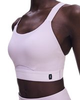 A close-up of a model wearing a light pink athletic sports bra with a wide underband and a small black On logo patch, paired with matching high-waisted bottoms against a white background. - On Performance Bra (Women's) - Foxglove