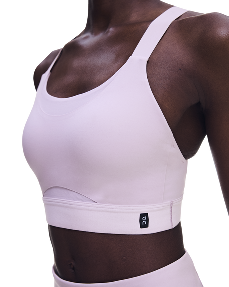 A close-up of a model wearing a light pink athletic sports bra with a wide underband and a small black On logo patch, paired with matching high-waisted bottoms against a white background. - On Performance Bra (Women's) - Foxglove