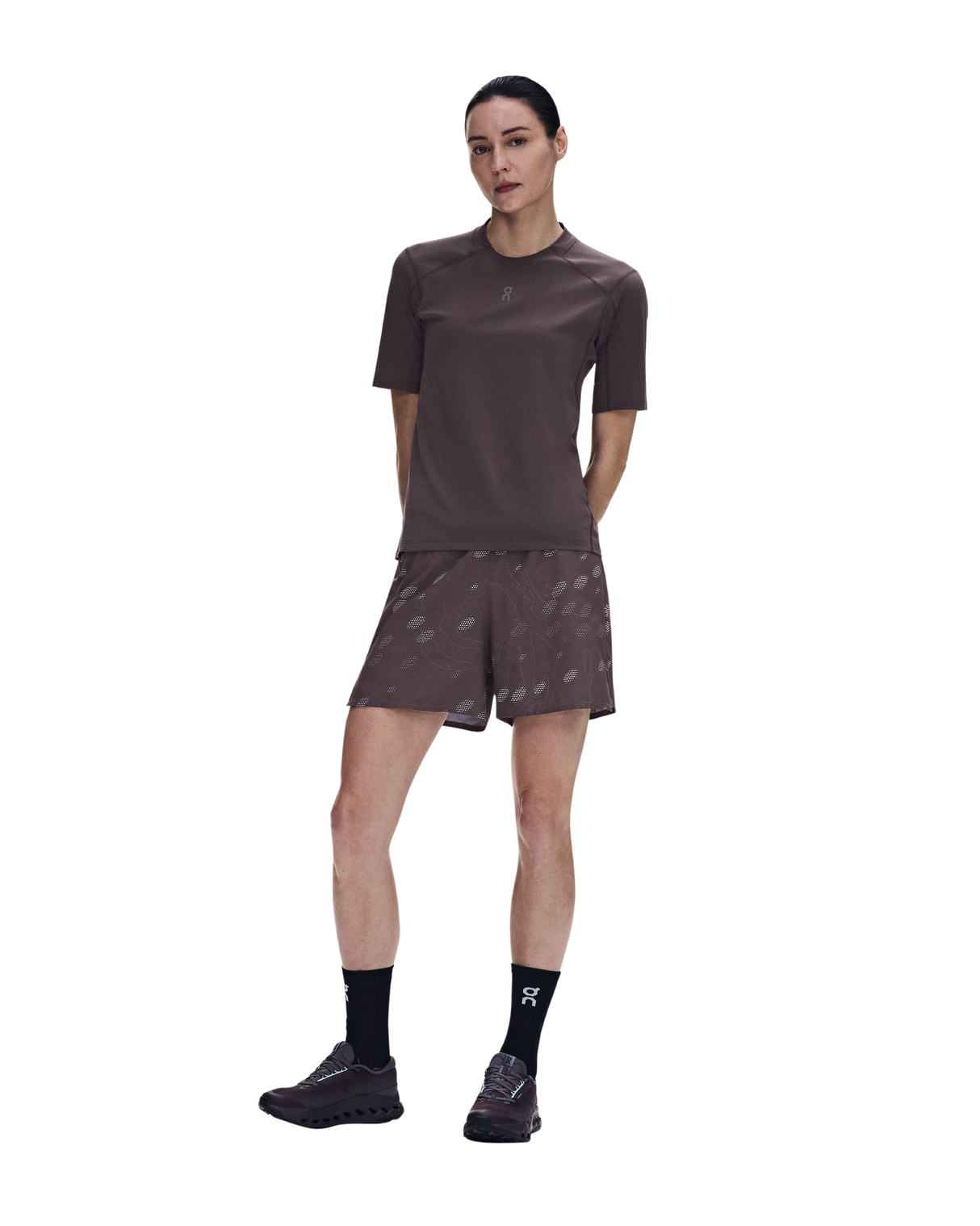 A woman modeling a matching athletic set consisting of a short-sleeve crewneck shirt and patterned shorts in a dark brown color, paired with black socks and trail running shoes, against a plain white background. - On Trail-T Solar (Women's) - Mulberry