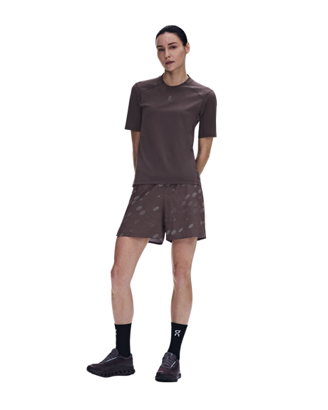 A woman modeling a matching athletic set consisting of a short-sleeve crewneck shirt and patterned shorts in a dark brown color, paired with black socks and trail running shoes, against a plain white background. - On Trail-T Solar (Women's) - Mulberry