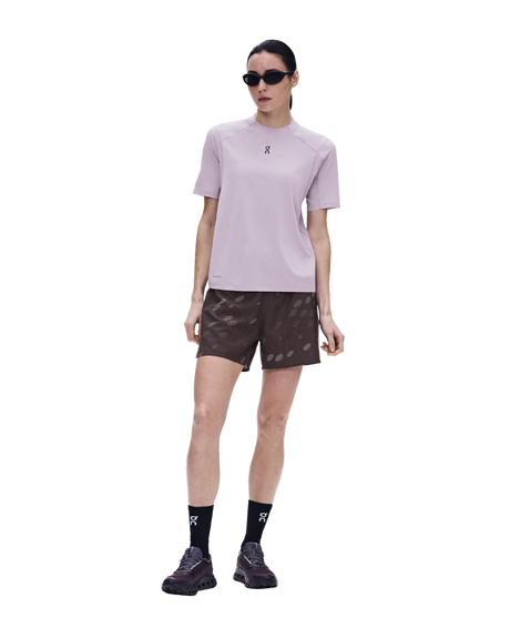 A model wearing a light purple athletic t-shirt, dark brown patterned shorts, black socks, and black running shoes, standing against a solid white background. - On Trail-T Solar (Women's) - Foxglove