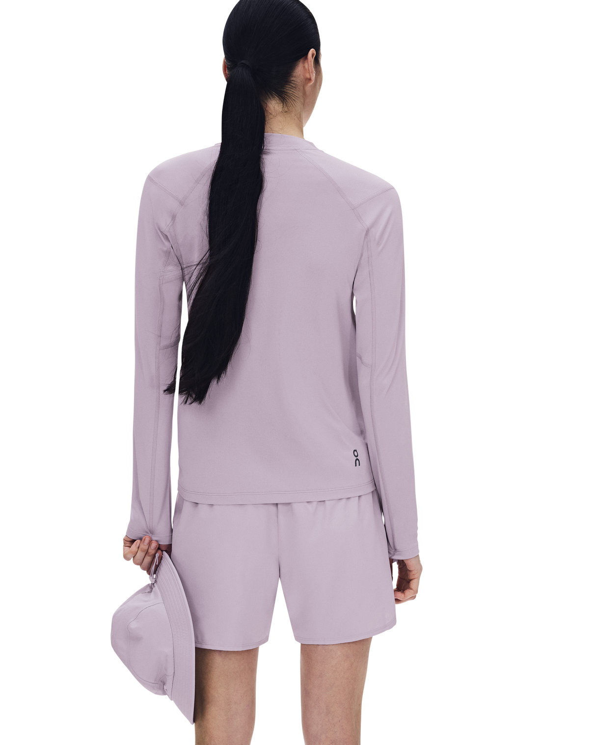 A woman shown from the back wearing a matching light lavender athletic set consisting of a long-sleeve top and shorts, with a matching cap in one hand, against a plain white background. - On Trail Long-T Solar (Women's) - Foxglove