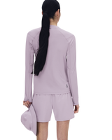 A woman shown from the back wearing a matching light lavender athletic set consisting of a long-sleeve top and shorts, with a matching cap in one hand, against a plain white background. - On Trail Long-T Solar (Women's) - Foxglove