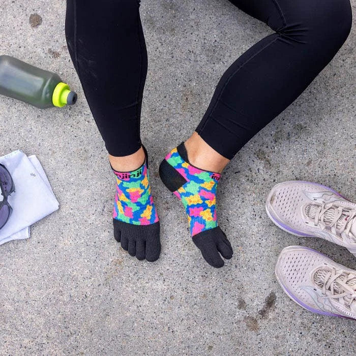 A person wearing colorful toe-separated athletic socks with black toes and heels, paired with black leggings and light gray running shoes, sitting on a concrete surface with a water bottle, towel, and sunglasses nearby. - Injinji Spectrum Run Lightweight No-Show Toesocks (Women's) - Gummy Bears