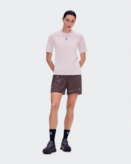 A model stands against a light gray studio background wearing a light pink athletic short-sleeve top, dark brown patterned shorts, black socks, and dark running shoes. The top has a small black logo on the left chest, and the socks and shoes show the same logo. - On Trail Shorts (Women's) - Mulberry