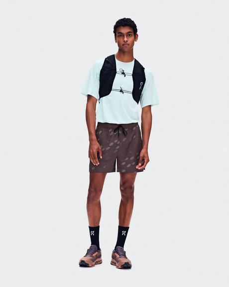 A male model stands against a light gray studio background wearing a white t-shirt, brown patterned shorts, black crew socks, trail running shoes, and a black hydration backpack. - On Trail Shorts (Men's) - Mulberry