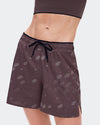 A close-up of a model wearing dark brown athletic shorts with a subtle white topographic pattern and a black drawstring waist, paired with a matching dark brown sports bra against a light gray background. - On Trail Shorts (Women's) - Mulberry