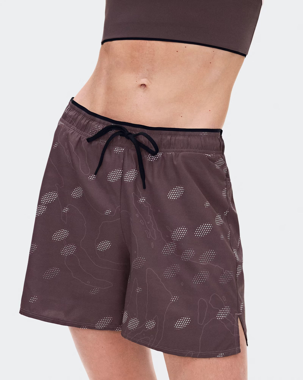 A close-up of a model wearing dark brown athletic shorts with a subtle white topographic pattern and a black drawstring waist, paired with a matching dark brown sports bra against a light gray background. - On Trail Shorts (Women's) - Mulberry