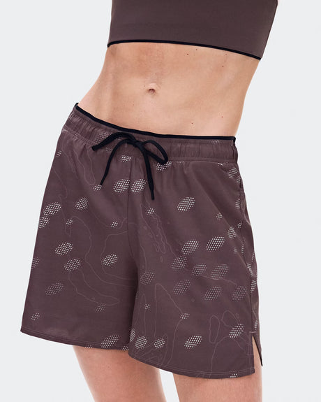 A close-up of a model wearing dark brown athletic shorts with a subtle white topographic pattern and a black drawstring waist, paired with a matching dark brown sports bra against a light gray background. - On Trail Shorts (Women's) - Mulberry