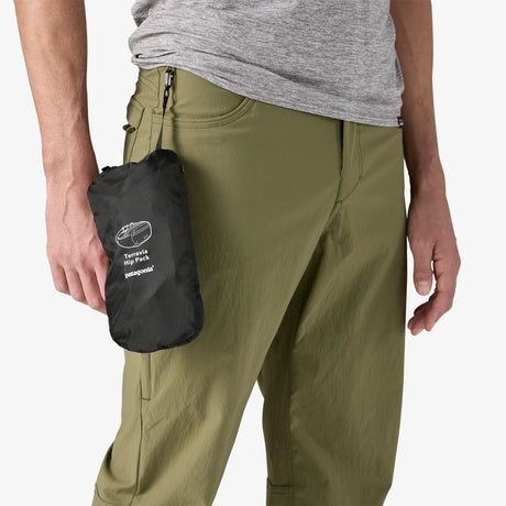 A close-up of a person wearing olive green pants and a heather gray shirt, holding a small black zip pouch with a white printed logo that reads 'Tarpack Tarpack Mini Pack' and 'Intrepid'. - Patagonia Terravia Hip Pack 4L - Black