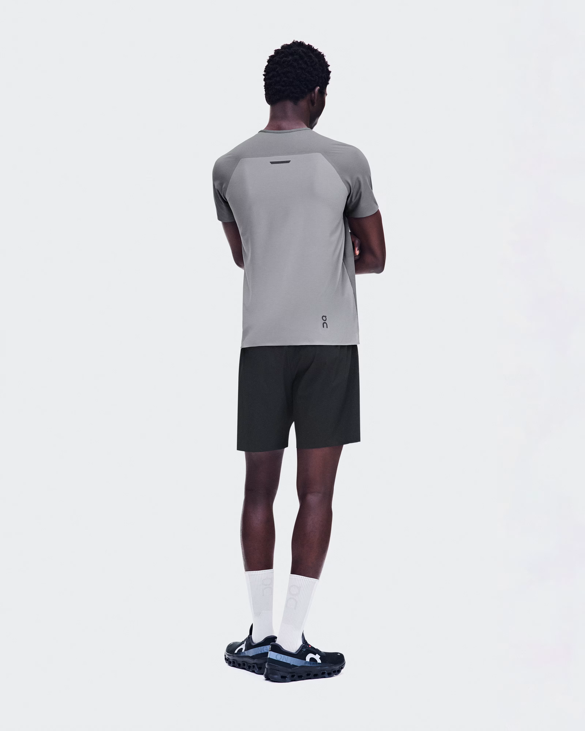 A man shown from the back wearing a light gray athletic t-shirt with dark gray raglan sleeves, black athletic shorts, white socks, and black running shoes with blue accents, against a solid light gray background. - On Performance-T (Men's) - Ember/Ore