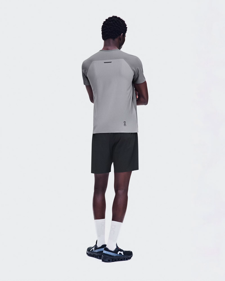 A man shown from the back wearing a light gray athletic t-shirt with dark gray raglan sleeves, black athletic shorts, white socks, and black running shoes with blue accents, against a solid light gray background. - On Performance-T (Men's) - Ember/Ore