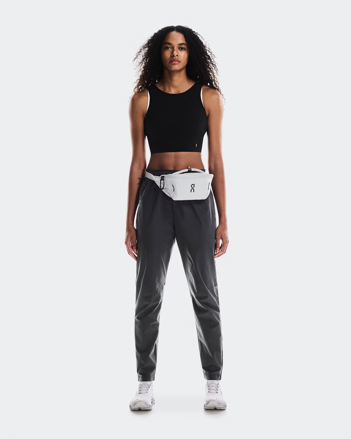 Woman wearing a black tank top and gray pants with a white waist bag on a white background - On Waist Pack 2L Lite - Glacier