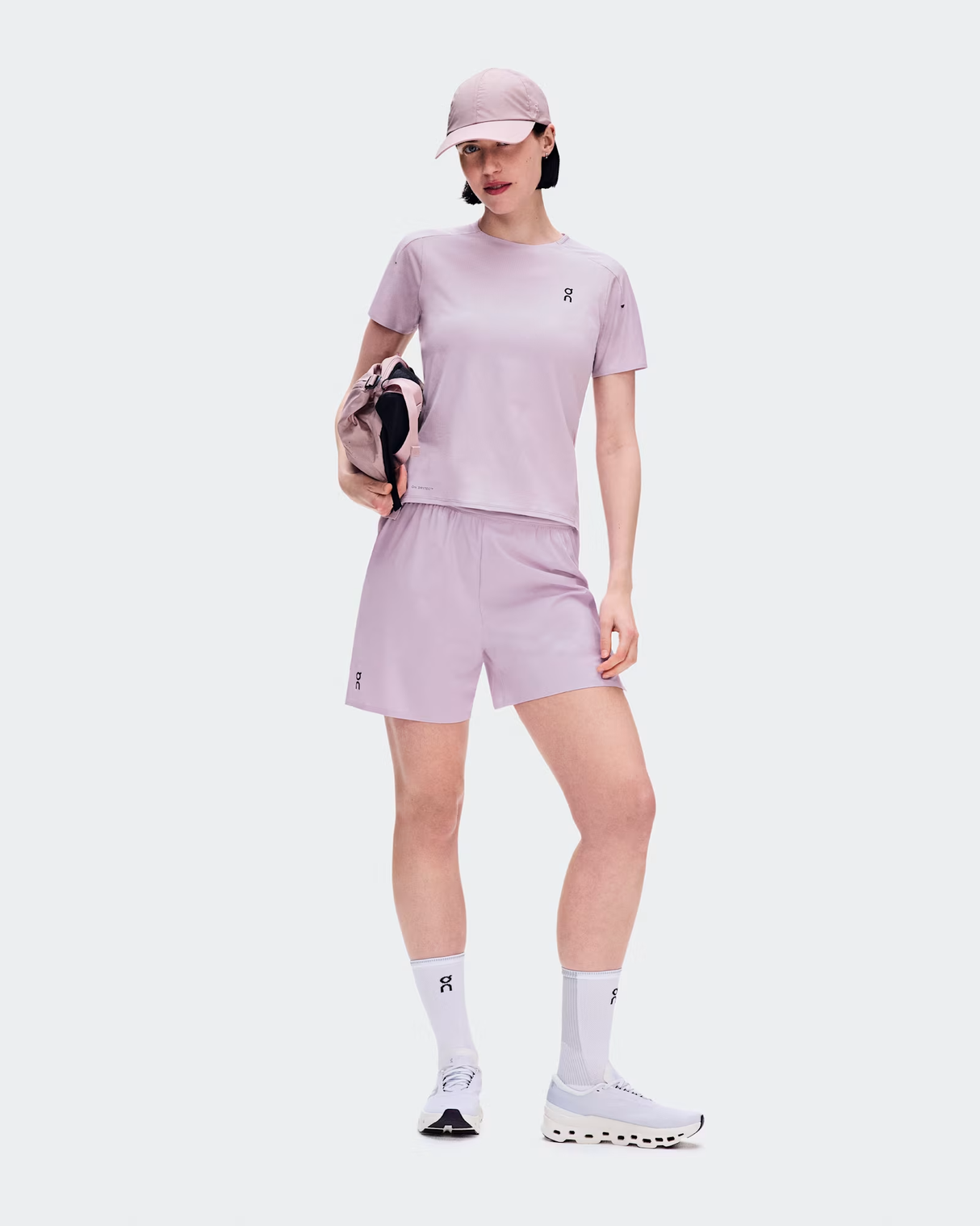 A model wearing a light purple athletic set consisting of a short-sleeve t-shirt, matching shorts, white socks, and white running shoes, holding a matching purple and black bag against a light gray background. - On Performance-T (Women's) - Foxglove/Orchid