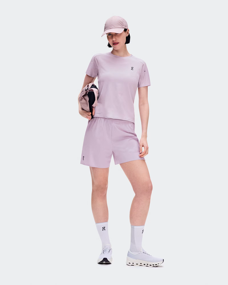 A model wearing a light purple athletic set consisting of a short-sleeve t-shirt, matching shorts, white socks, and white running shoes, holding a matching purple and black bag against a light gray background. - On Performance-T (Women's) - Foxglove/Orchid
