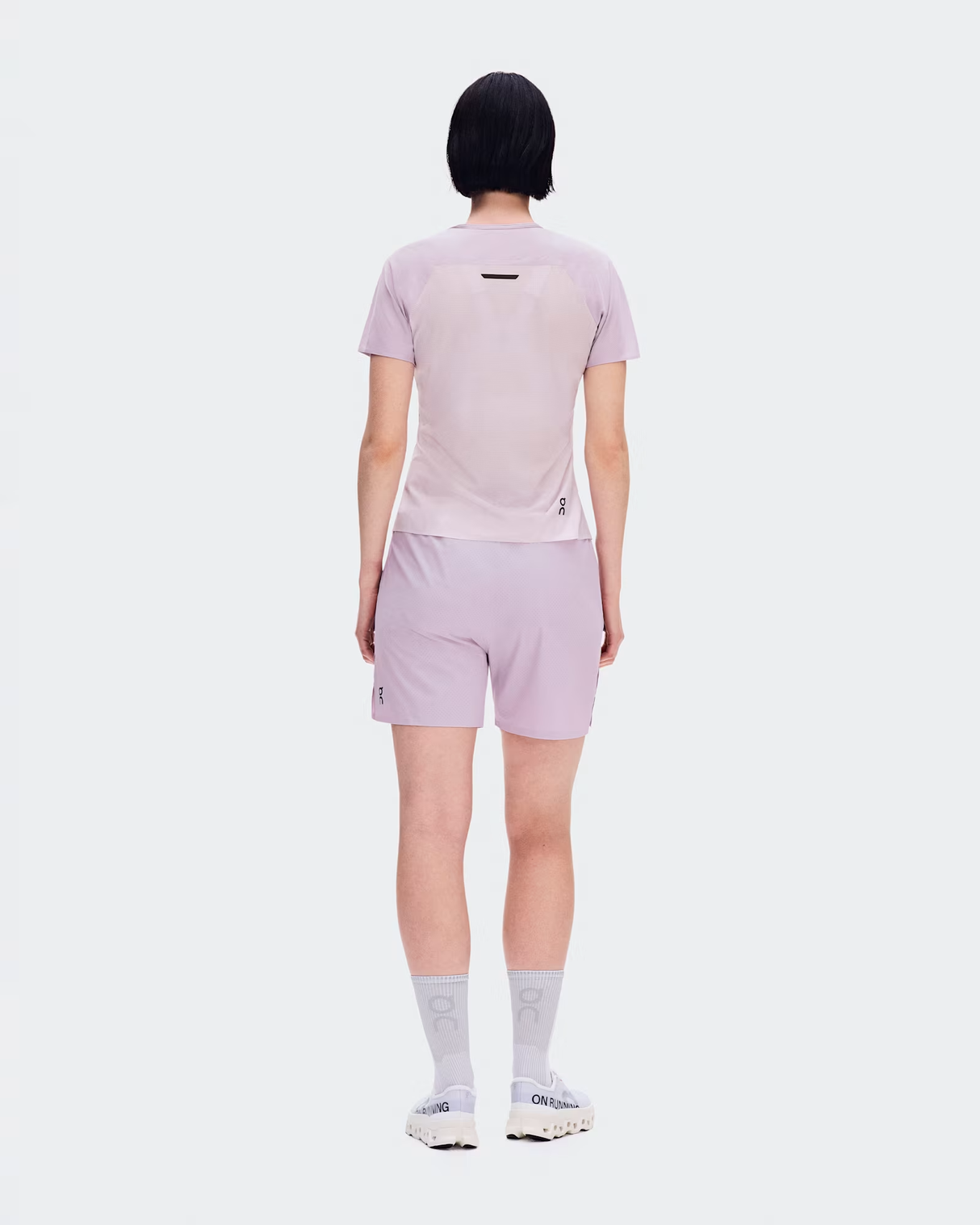 A model viewed from the back wearing a light pink athletic t-shirt and matching light pink shorts, paired with white socks and white sneakers, against a solid white background. - On Performance-T (Women's) - Foxglove/Orchid