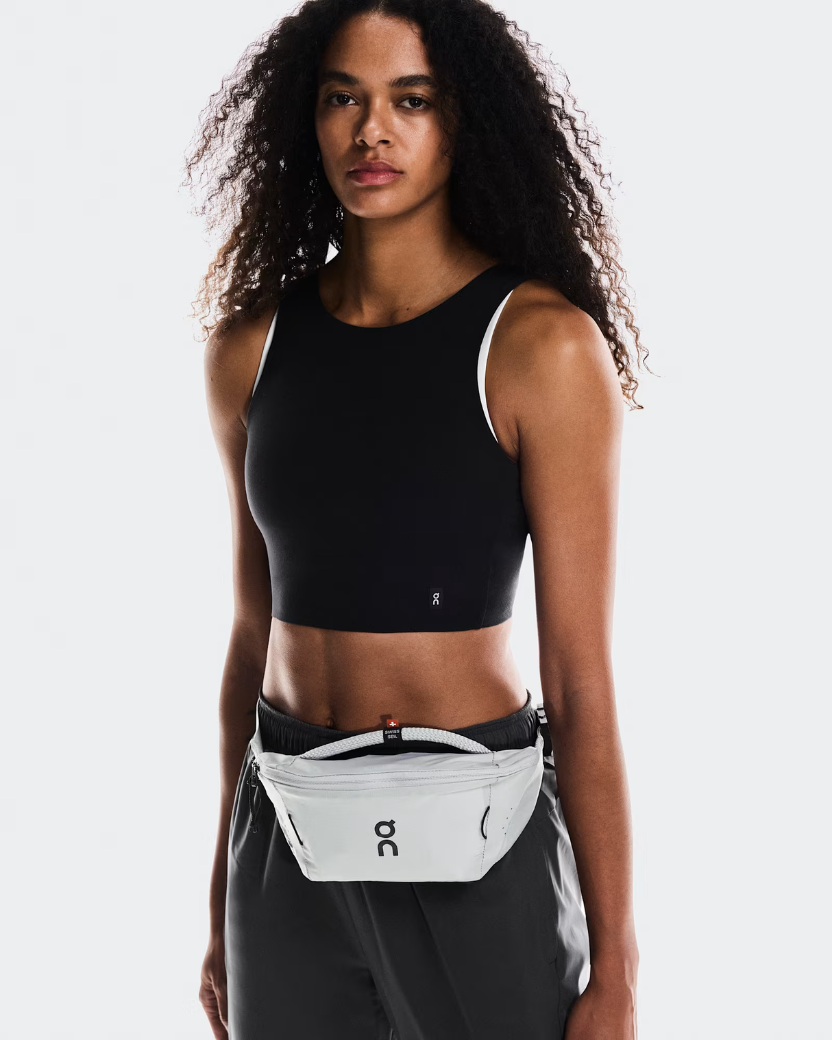 Woman wearing a black sports bra and gray leggings with a white waist bag on a light gray background - On Waist Pack 2L Lite - Glacier