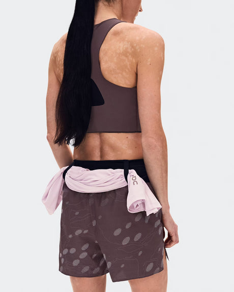 Rear view of a woman wearing a dark brown athletic sports bra and matching patterned shorts, with a light pink shirt tied around her waist, against a light gray studio background. - On Trail Shorts (Women's) - Mulberry