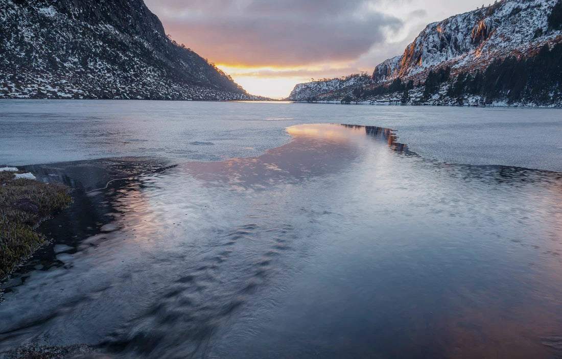A scenic winter landscape with a frozen or partially frozen lake between snow-dusted mountains, under a sunset sky reflected in the water. - Rob Blakers Tasmanian Landscapes Calendar