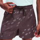 A close-up of a person wearing dark brown swim trunks with a white topographic dot pattern and a black drawstring, paired with a light mint green shirt. - On Trail Shorts (Men's) - Mulberry