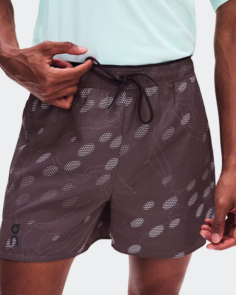 A close-up of a person wearing dark brown swim trunks with a white topographic dot pattern and a black drawstring, paired with a light mint green shirt. - On Trail Shorts (Men's) - Mulberry