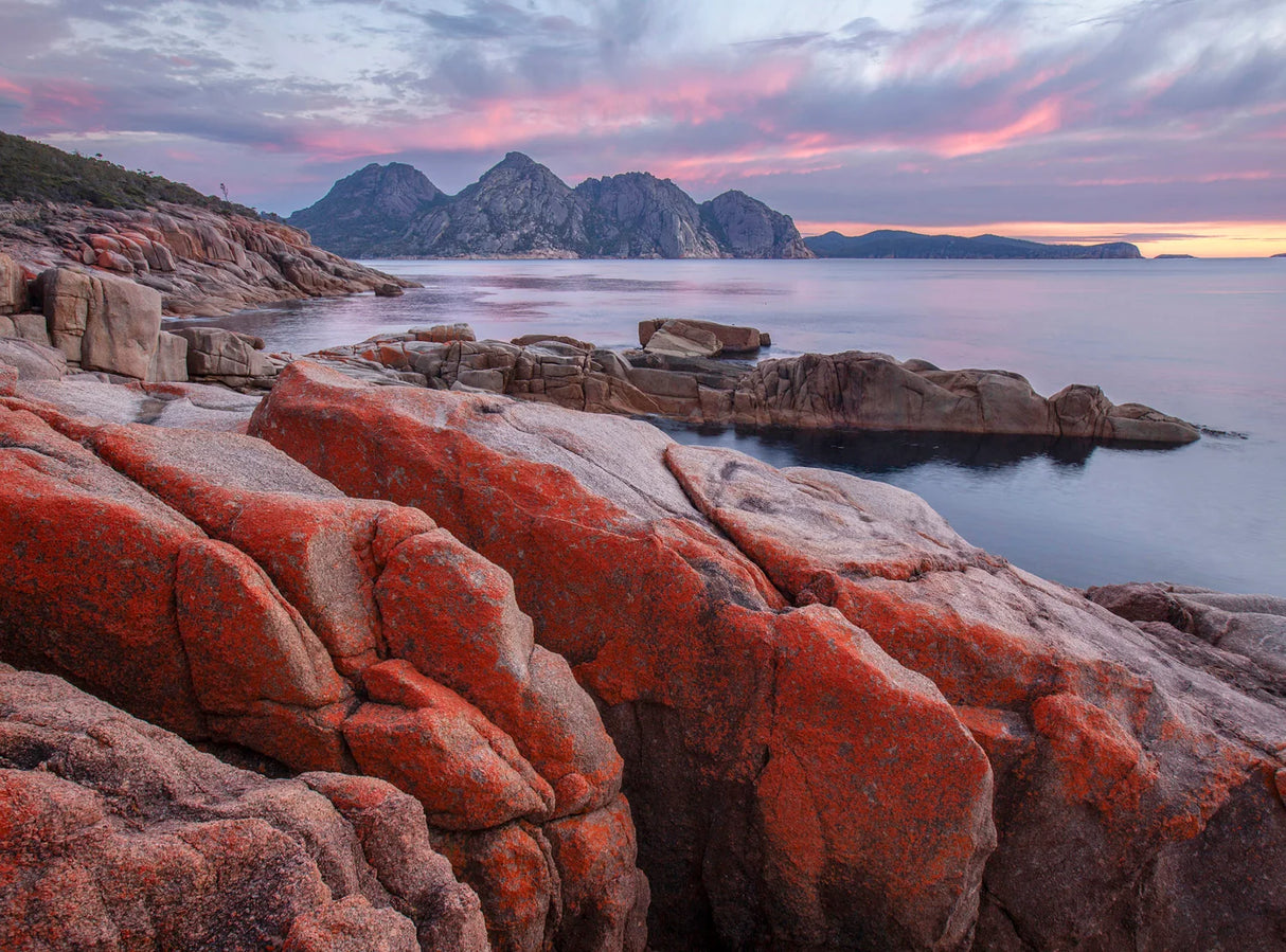 A scenic coastal landscape with large orange-red rocks in the foreground, calm water, distant rocky mountains, and a pink and purple sunset sky. - Rob Blakers Wild Tasmania 2027 Large Calendar