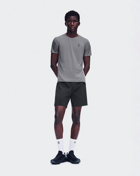 A male model stands against a light gray studio background wearing a gray short-sleeve athletic T-shirt with a small black logo on the left chest, dark athletic shorts, white socks, and black sneakers. - On Performance-T (Men's) - Ember/Ore