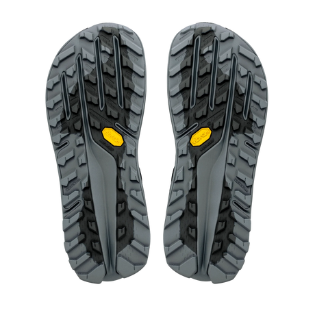 A pair of rugged hiking shoe soles shown from the bottom, featuring a dark gray and black tread pattern with small yellow Vibram logos. - Altra Olympus 7 Shoes (Women's) - Black/Silver/Castlerock