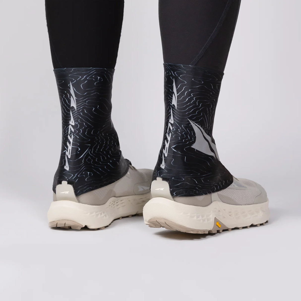 Person wearing black patterned socks with white shoes on a light gray background - Altra Trail Gaiters - Black/Silver/Grey