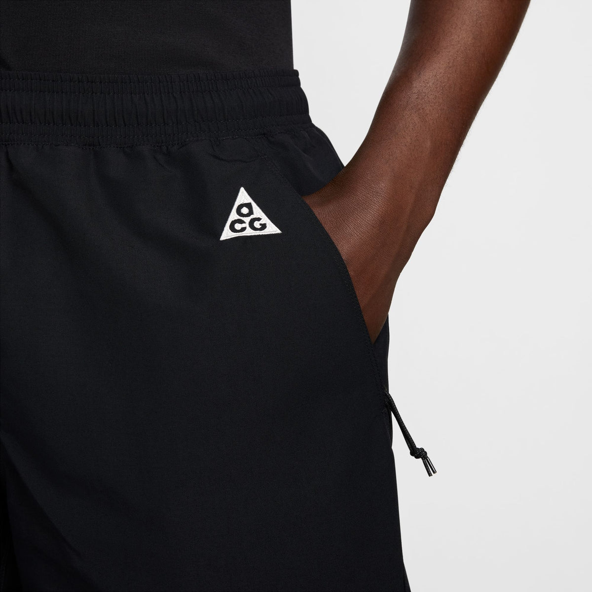 Close-up of a person wearing black athletic shorts with a white triangular ACG logo on the left hip, shown against a light gray studio background. - Nike ACG Reservoir Goat Shorts (Men's) - Black/Anthracite/Summit White