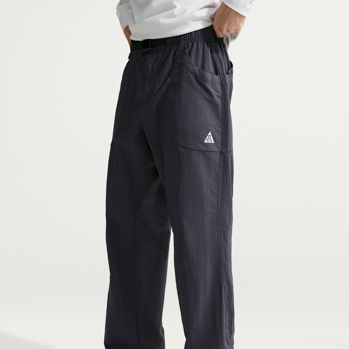 A person wearing dark charcoal gray technical cargo pants with a small white ACG logo on the pocket, paired with a white shirt, shown from the waist down against a light gray studio background. - Nike ACG Dolomiti Pants (Men's) - Gridiron/Summit White