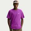 A man wearing a bright purple short-sleeve crewneck t-shirt with a small white geometric logo on the left chest, paired with brown pants and an orange and white cap with sunglasses. - Nike ACG Dri-FIT T-Shirt (Men's) - Bold Berry/Summit White