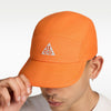 A close-up of a person wearing an orange cap with a white embroidered triangle logo featuring the letters 'a', 'c', and 'g'. The person is holding the brim of the cap with their hand against a clean white background. - Nike ACG Fly Unstructured Cap - Safety Orange/Safety Orange
