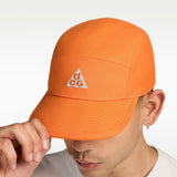 A close-up of a person wearing an orange cap with a white embroidered triangle logo featuring the letters 'a', 'c', and 'g'. The person is holding the brim of the cap with their hand against a clean white background. - Nike ACG Fly Unstructured Cap - Safety Orange/Safety Orange