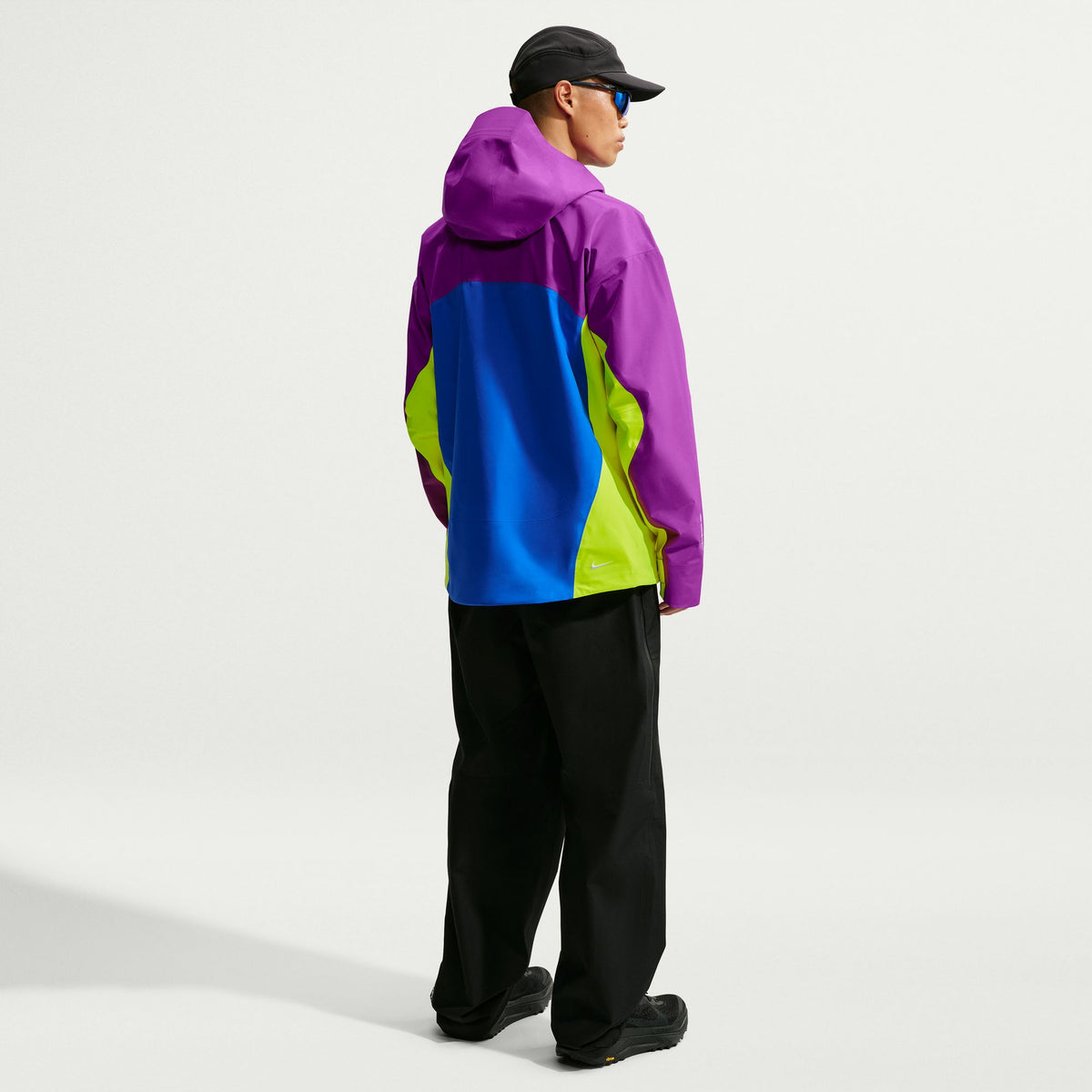 A model from the back wearing a color-blocked hooded jacket with purple, blue, and lime green panels, black pants, and a black cap, photographed against a clean white studio background. - Nike ACG Misery Ridge Storm-FIT ADV GORE-TEX Jacket (Men's) - Bold Berry/Hyper Royal/Lemon Venom