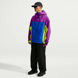 A model wearing a color-blocked hooded jacket in purple, blue, and neon yellow, paired with black pants, black shoes, and a black cap, standing against a light gray studio background. - Nike ACG Misery Ridge Storm-FIT ADV GORE-TEX Jacket (Men's) - Bold Berry/Hyper Royal/Lemon Venom